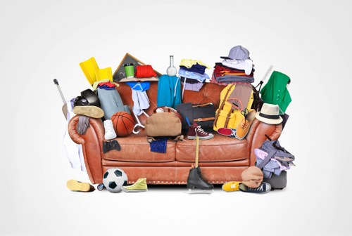A beige fabric sofa is densely covered with a variety of discarded items, including clothing, sports equipment such as a basketball and a soccer ball, and miscellaneous household objects. The clothing comprises shirts, pants, and other textiles in different colours and fabrics, some folded while others draped over the sofa edges. There is a brown leather bag, a pair of sneakers, a watering can, and plastic containers among the clutter. A hockey stick and a broom are partially visible, leaning against the sofa on the left. The scene is set against a plain white background, highlighting the accumulated rubbish, which appears to have been gathered for removal or disposal, consistent with the services offered by Garden Clearance Gunnersbury. The arrangement suggests an informal collection of items typical of a home clearance, with no specific order or organization, emphasizing the need for professional rubbish removal. This detailed description aims to assist accessibility, conveying the visual scope of the clutter and its setting, and subtly supports local SEO through contextual references to rubbish and clearance activities in the Gunnersbury area.