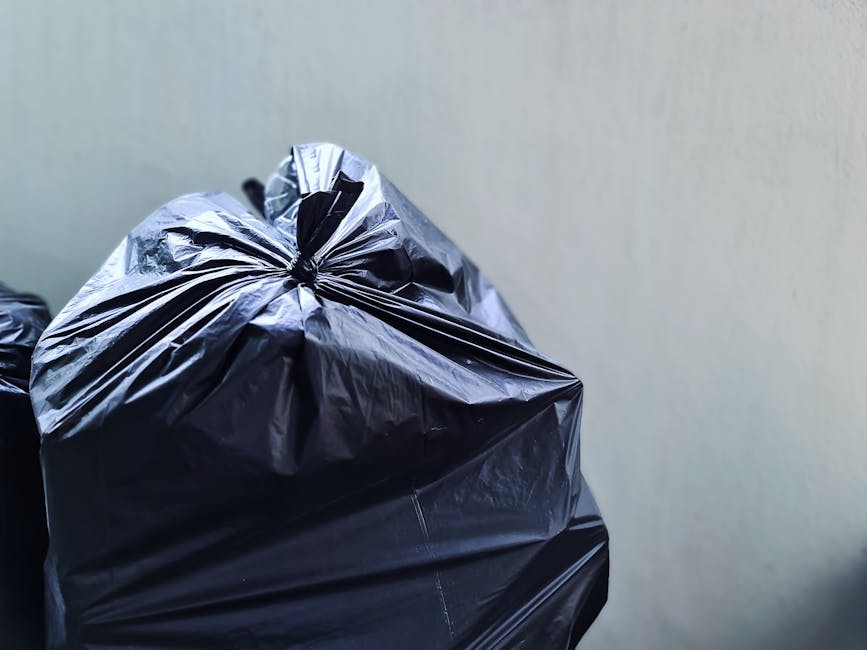 A large pile of mixed rubbish and waste materials is overflowing from a metal recycling bin situated on a paved sidewalk in an urban area. The waste includes crumpled cardboard boxes, plastic bags, lo